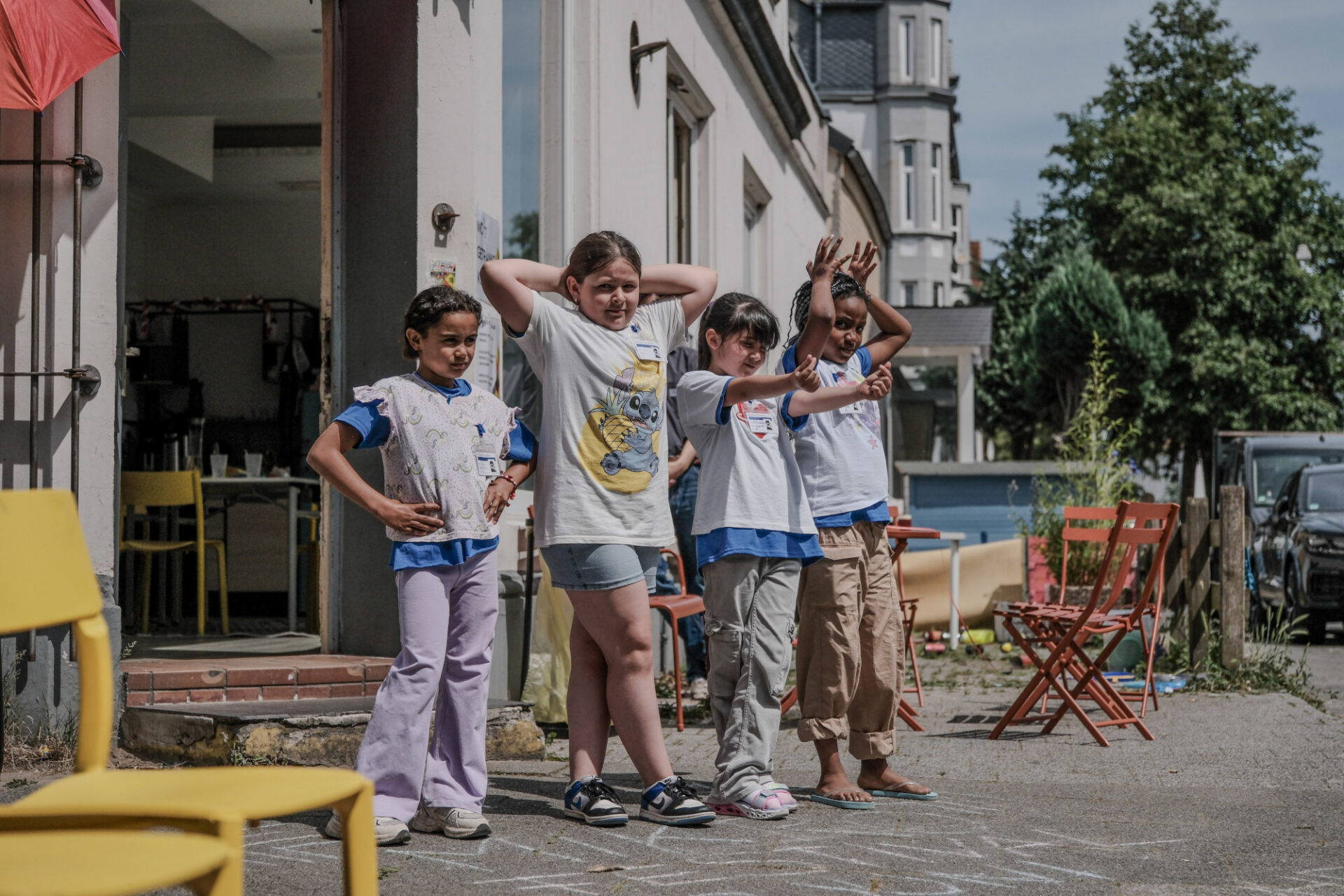 Vor einem Haus mit geöffneter Tür befinden sich vier Jugendliche sommerlich bekleidet in Tanzpose. Links und rechts davon sind Stühle aufgestellt, im Hintergrund befinden sich weitere Häuser, Bäume und parkende Autos.