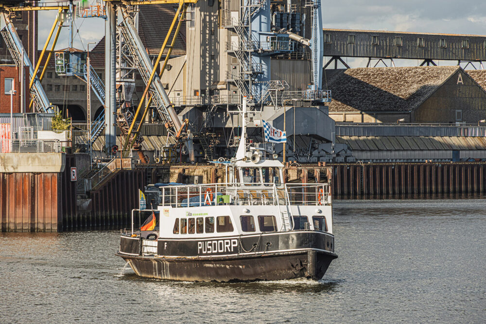 Eine kleine schwarz‑weiße Fähre mit dem Namen „PUSDORP“ fährt durch ein Hafenbecken. Am Heck weht eine Flagge. Im Hintergrund stehen große Industriehallen, Kräne und metallene Hafenanlagen. Der Himmel ist dicht bewölkt, das Wasser wirkt ruhig.