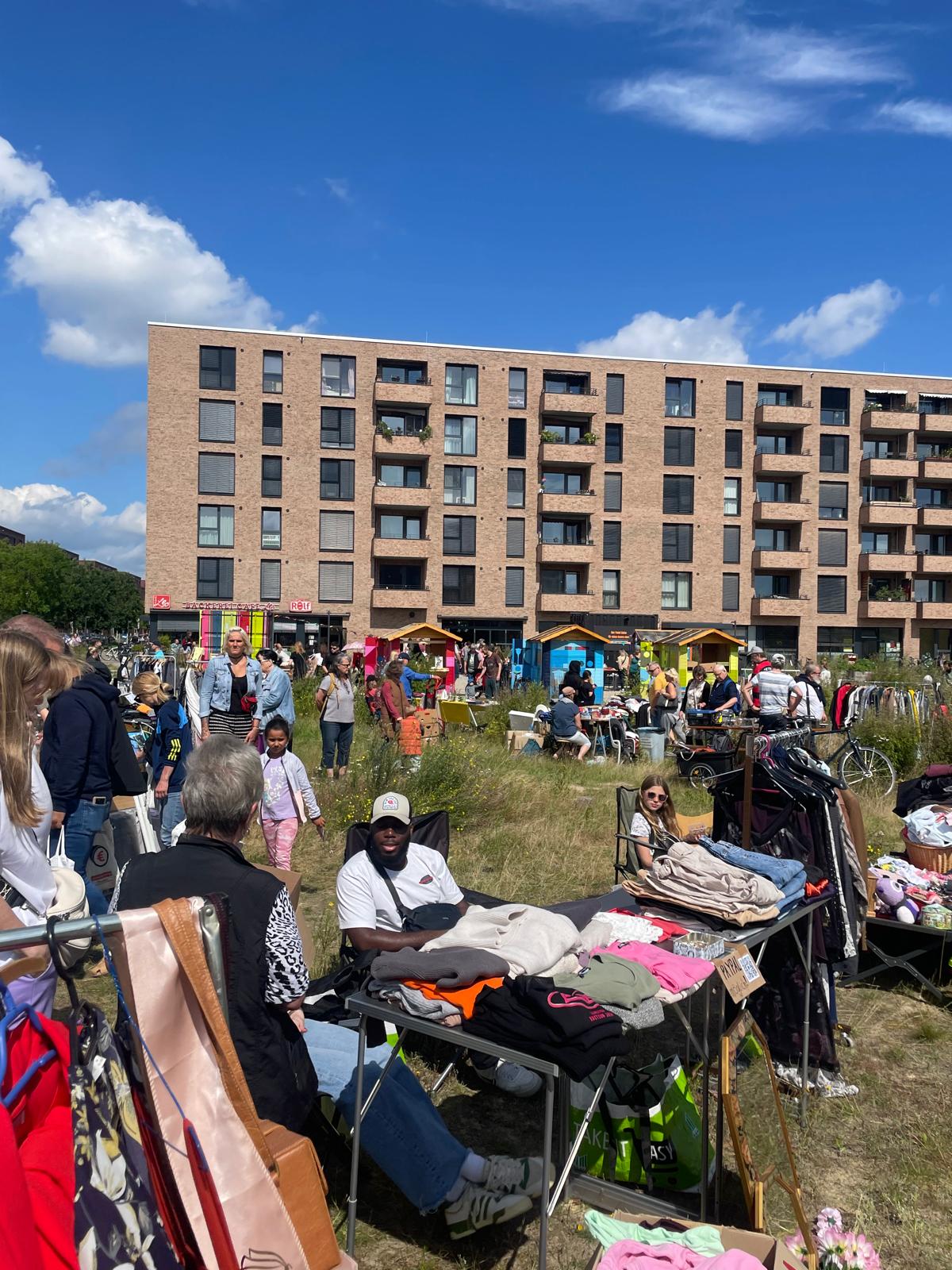 Eine Flohmarktsituation bei bestem Sonnenschein auf einer Wiese. Im Hintergrund ist ein Hochhaus zu sehen und ein blauer Himmel. 