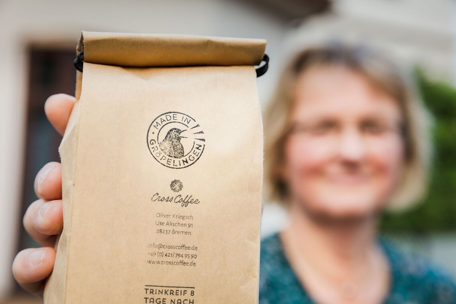 Eine Person, die nur verschwommen zu sehen ist, hält eine braune Packung in der Hand und zeigt diese ins Bild. Auf der Packung steht Made in Gröpelingen, Cross Coffee.